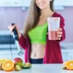 Healthy woman in sportswear preparing a fresh protein shake for weight loss, surrounded by organic fruits, in a modern kitchen.