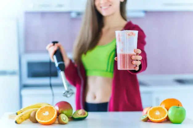 Healthy woman in sportswear preparing a fresh protein shake for weight loss, surrounded by organic fruits, in a modern kitchen.